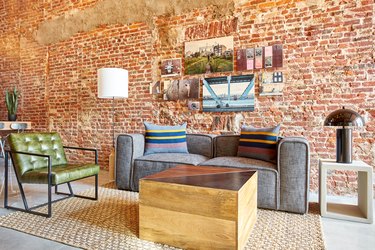 Industrial loft living room with gray couch, wood block coffee table, green leather armchair, chrome lamp, floor lamp, jute rug, brick wall, art, side table.